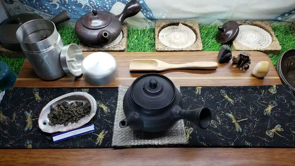 A still life of a traditional tea setup featuring multiple teapots, a wooden scoop, a metal tea canister with lid, dried tea…
