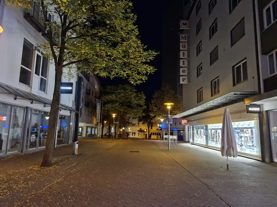 A nighttime street scene in a European city with illuminated storefronts, including 'SCHMID' and 'RABE', and a tall building…