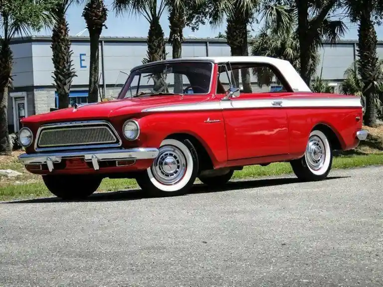A red and white 1960s American convertible car is parked on asphalt with palm trees in the background. The car has a white…