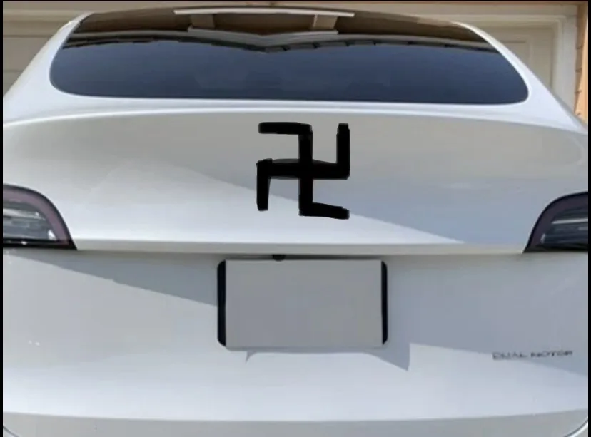 The rear of a white Tesla Model 3 is shown with a black swastika symbol digitally overlaid on the trunk, directly above the…