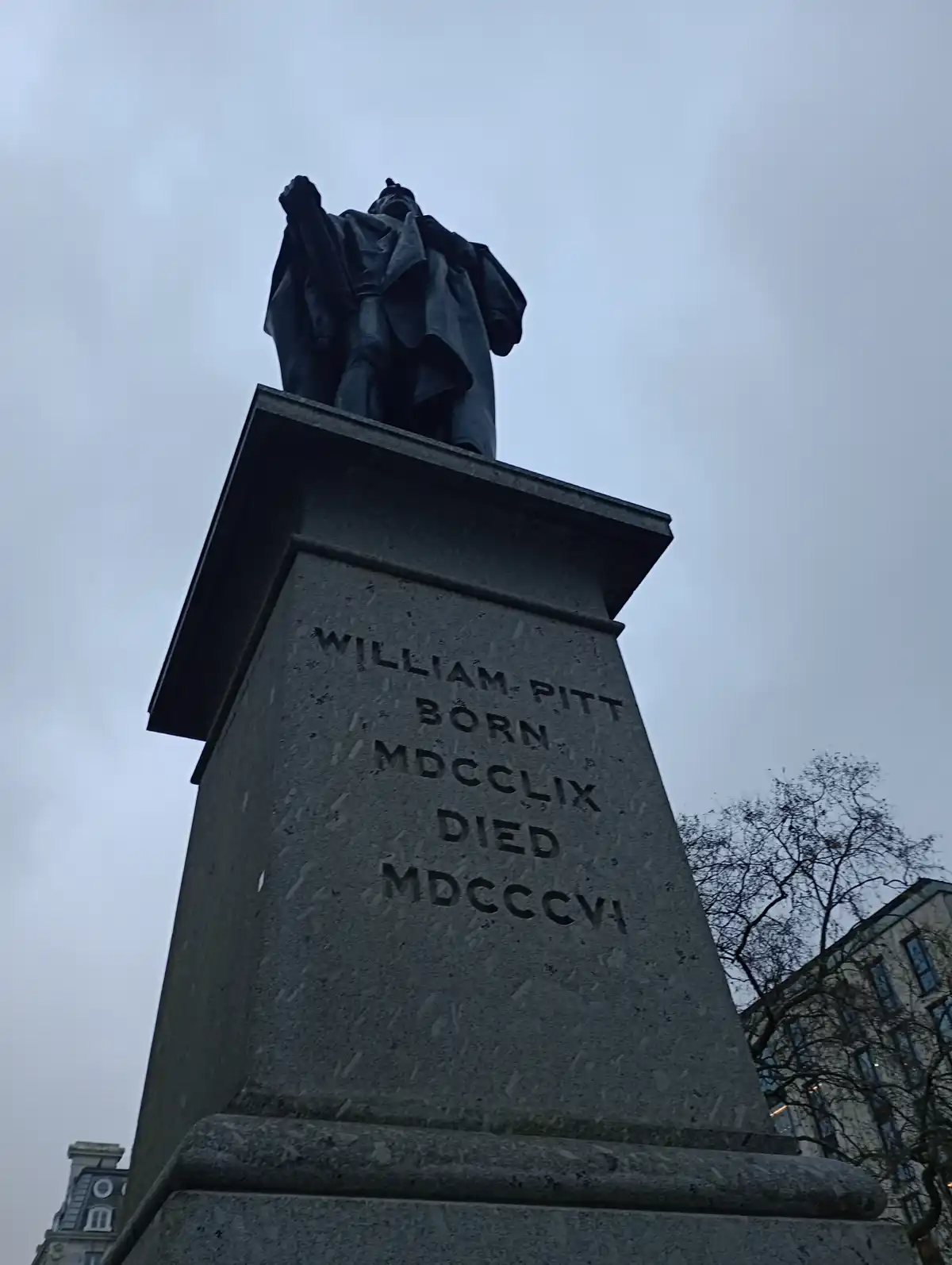A low-angle photo of a dark statue atop a stone pedestal under an overcast sky, with engraved text reading 'WILLIAM PITT…