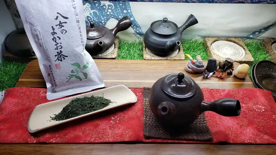 A still life of a traditional Japanese tea setup featuring a bag of '八女のよかお茶' (Yayoi's Good Tea) with Japanese text, loose…