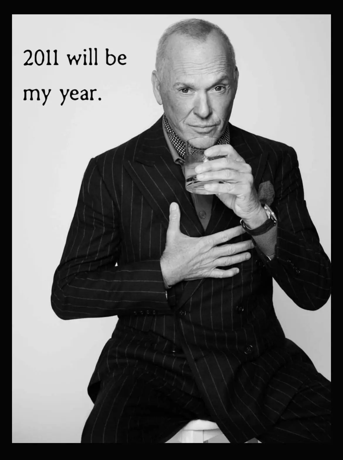 A black and white photo shows a man in a pinstripe suit holding a glass to his lips while placing his other hand over his…