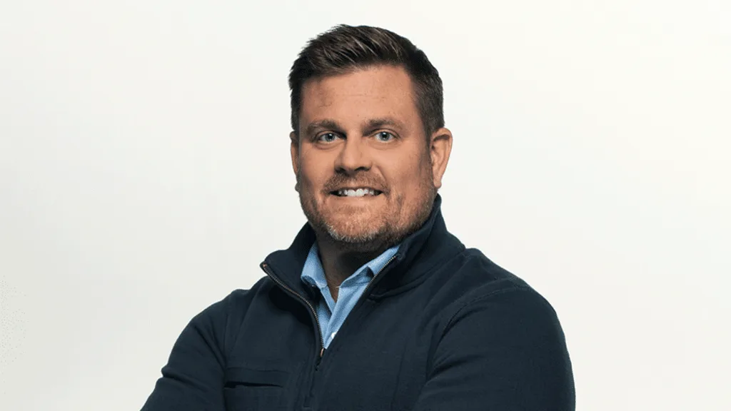 a man with short brown hair and a beard, wearing a dark blue zip-up jacket over a light blue collared shirt, smiles slightly…