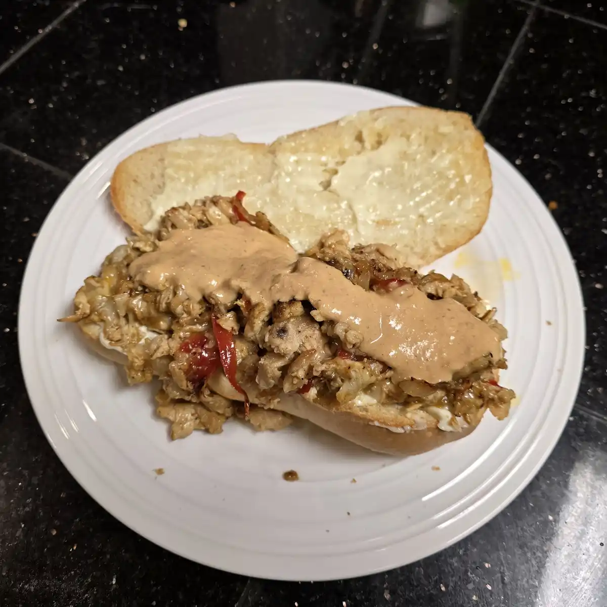 A close-up photo shows a sandwich on a white plate, with shredded meat, red peppers, and a thick brown sauce on the bottom…