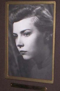 A black-and-white portrait of a woman with styled hair and a serious expression, framed in gold and mounted on a dark wall…