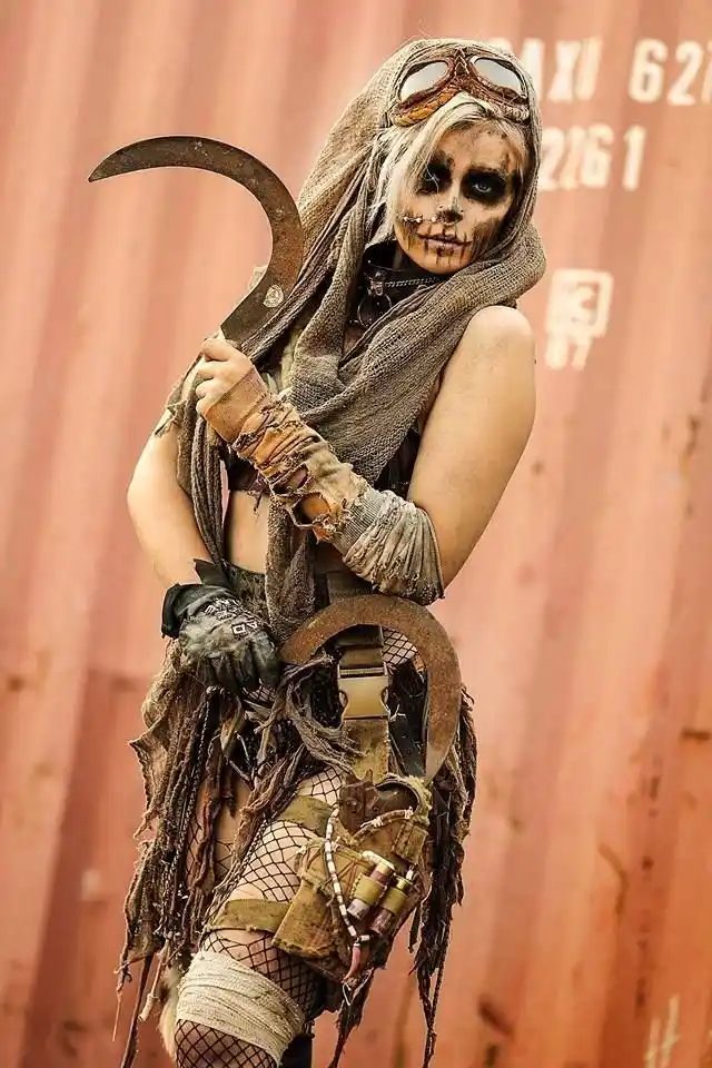 A person dressed as a grim reaper or post-apocalyptic warrior, wearing skull makeup, goggles, a tattered hood, and holding a…