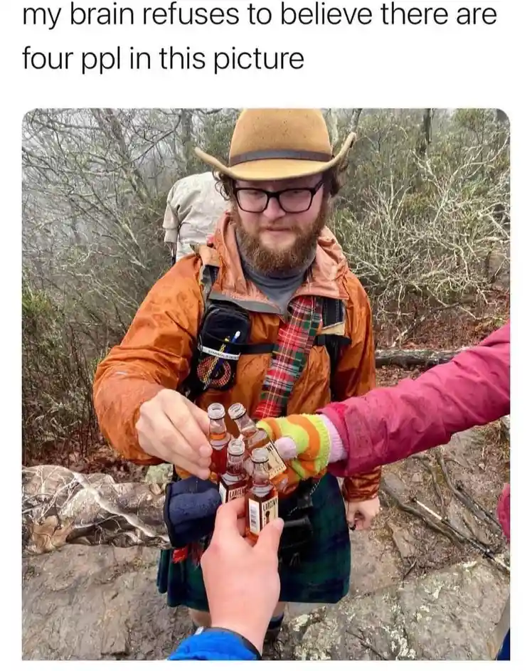 A man in an orange jacket, kilt, and cowboy hat holds several miniature liquor bottles while standing on a rocky trail. Two…