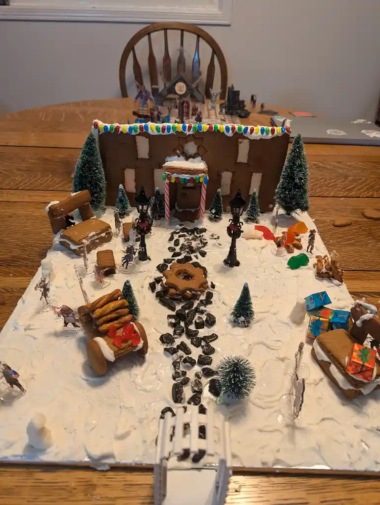 A detailed gingerbread house model sits on a wooden table, surrounded by white icing 'snow', miniature trees, candy…