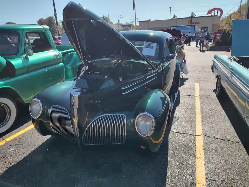 A dark green vintage car with its hood open is parked in a lot surrounded by other classic cars, including a teal pickup…