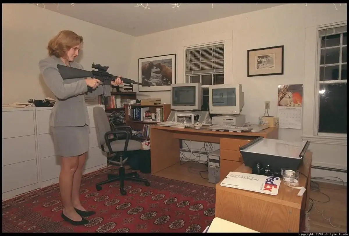 A woman in a grey business suit stands in a cluttered 1990s office holding an assault rifle, aiming it toward the viewer…