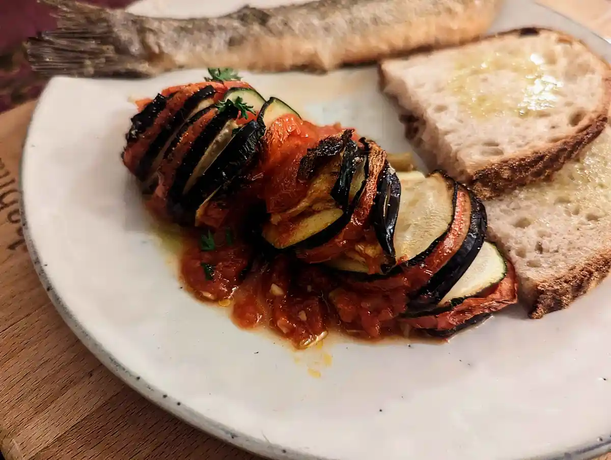 A plate of food shows a whole fried fish with its tail visible, alongside roasted eggplant and tomato slices in sauce, and…