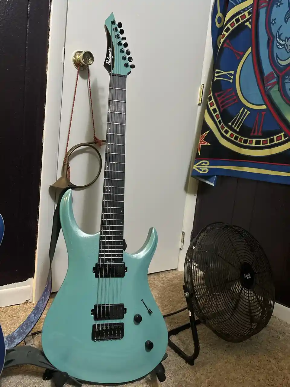 A light blue electric guitar with a black neck and headstock is propped up against a white door, its strap hanging down. To…