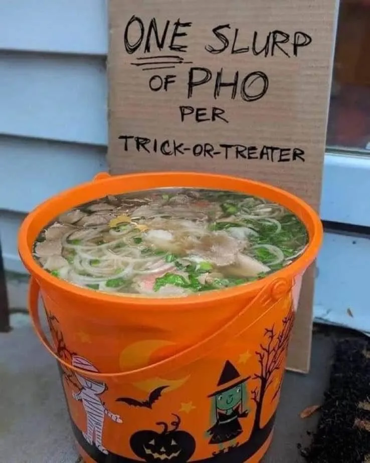 An orange Halloween bucket, decorated with pumpkins, bats, and cartoon witches, is filled with pho soup. Behind it, a…