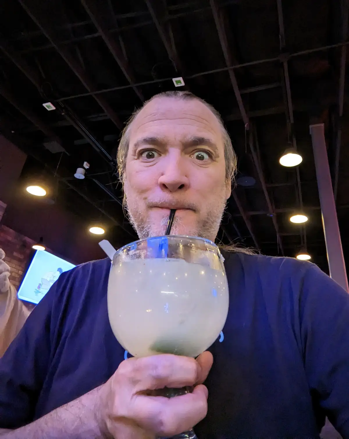 A man with a beard and long hair, wearing a dark blue shirt, holds a large glass of pale-colored cocktail with a black straw…