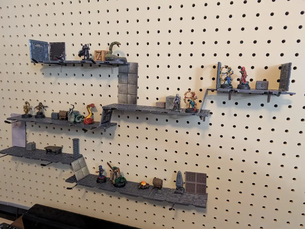 A beige pegboard wall has multiple gray, modular shelves attached, each holding miniature fantasy figures, terrain pieces…