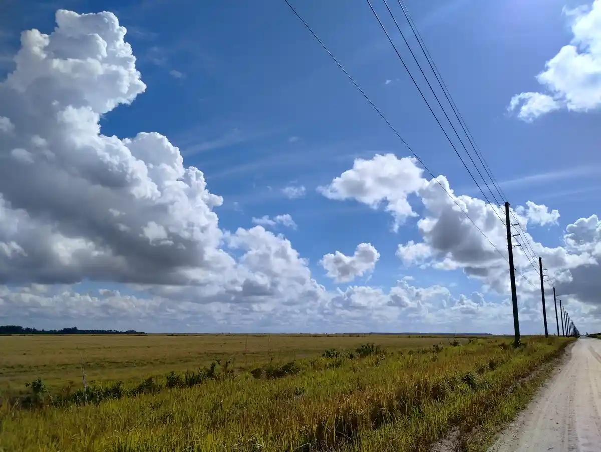 A wide-angle photo shows a dirt road stretching into the distance through a flat, grassy field under a bright blue sky with…