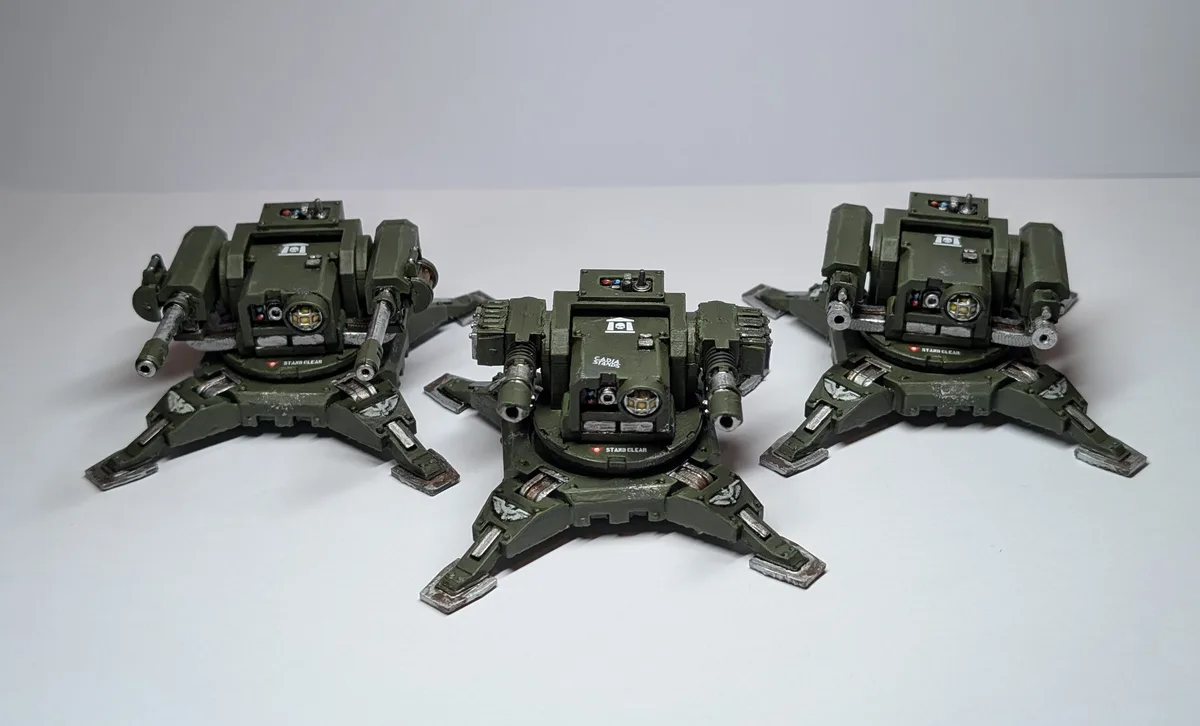 Three green, heavily armed, spider-like war machines are arranged in a triangle on a white surface. Each has multiple…