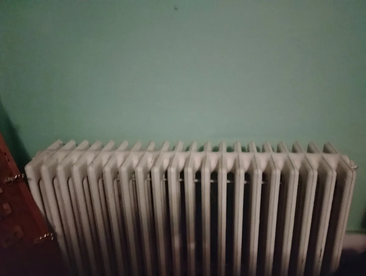 A close-up photo shows a white, vertical-panel radiator against a teal wall, with a wooden door partially visible to the…