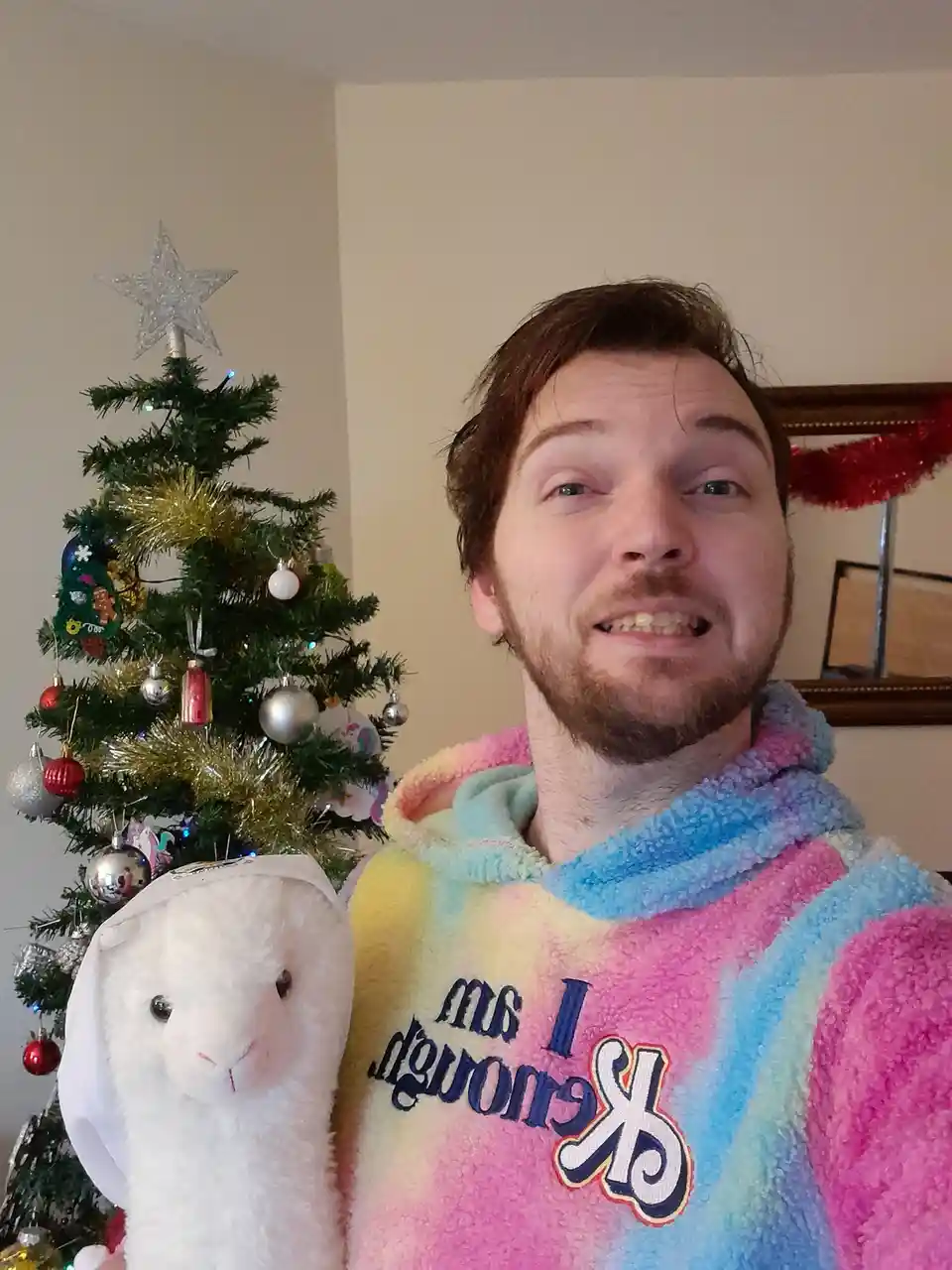 A man with a beard and messy hair wears a rainbow-colored, fluffy hoodie with the text 'I saw you' and a stylized 'K' logo…