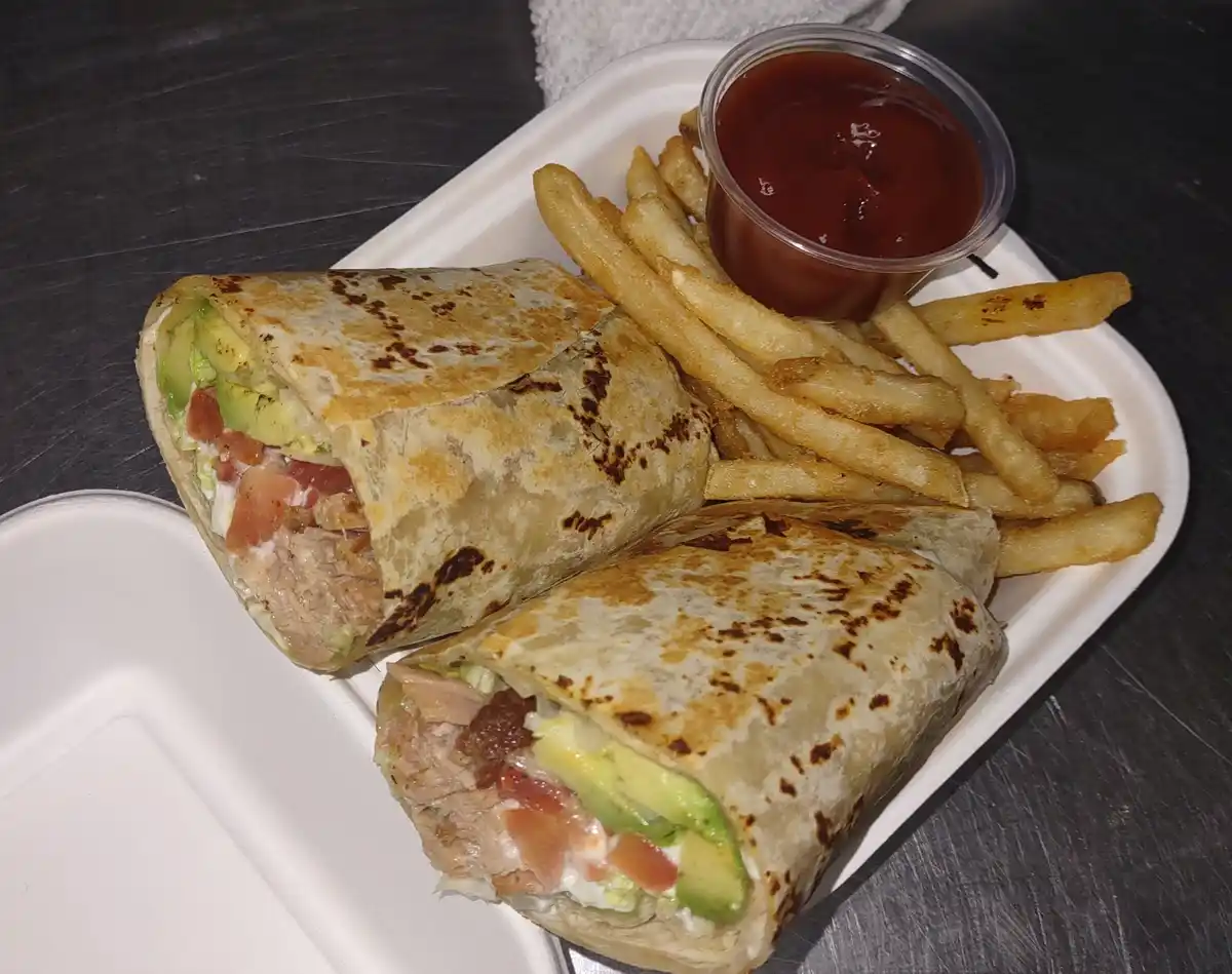 A white styrofoam container holds a burrito cut in half, revealing fillings of meat, avocado, tomato, and sauce, alongside…