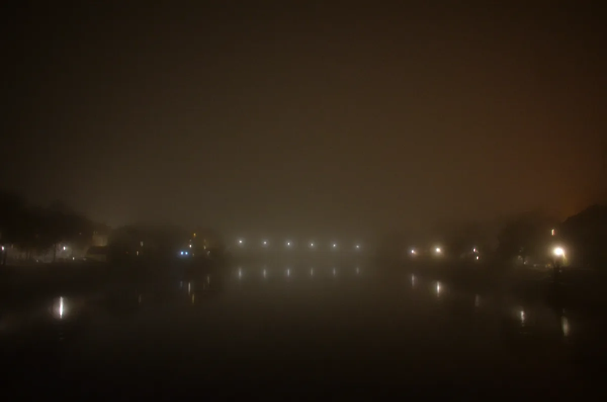 A dark, foggy night scene with distant streetlights and their reflections on wet pavement, creating a mysterious, blurry…