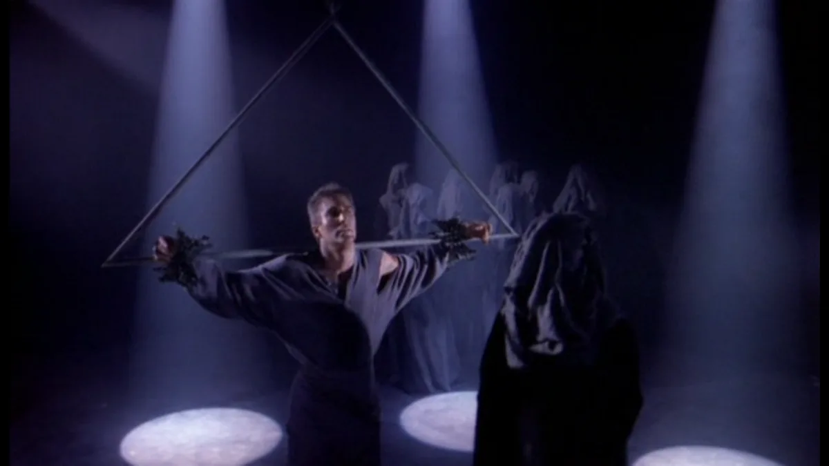 A man in a dark robe stands with arms outstretched, holding a long staff that forms the base of an inverted triangle, while…