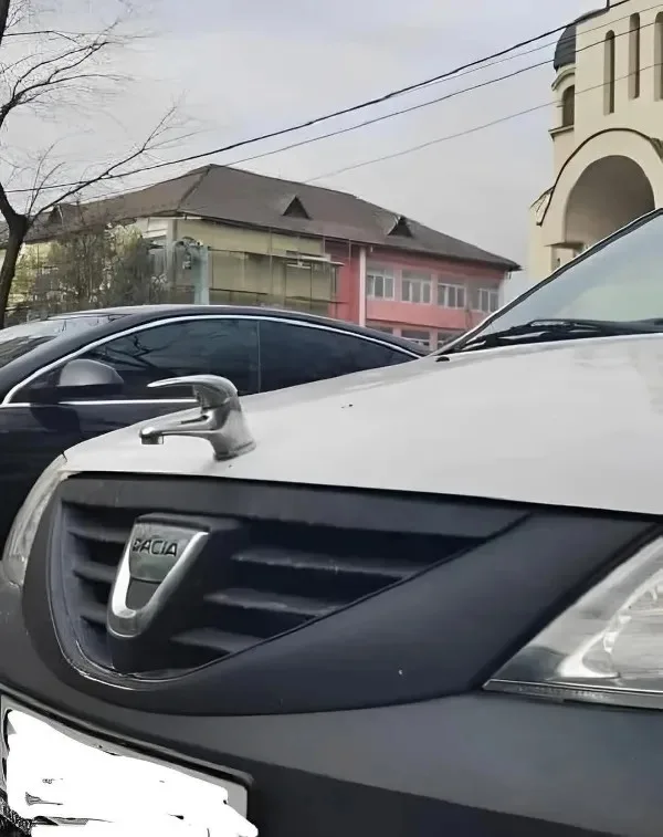 A silver faucet is visibly mounted on the hood of a white Dacia car, directly above the grille. The car is parked outdoors…