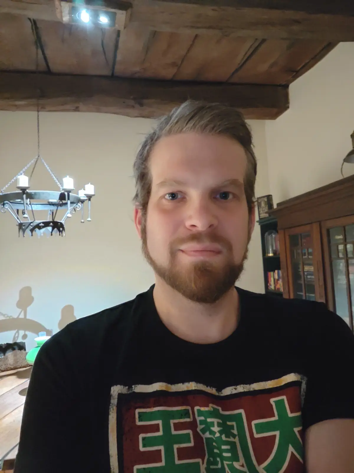 A man with a beard and short hair wearing a black t-shirt with large green Chinese characters on it stands in a room with…