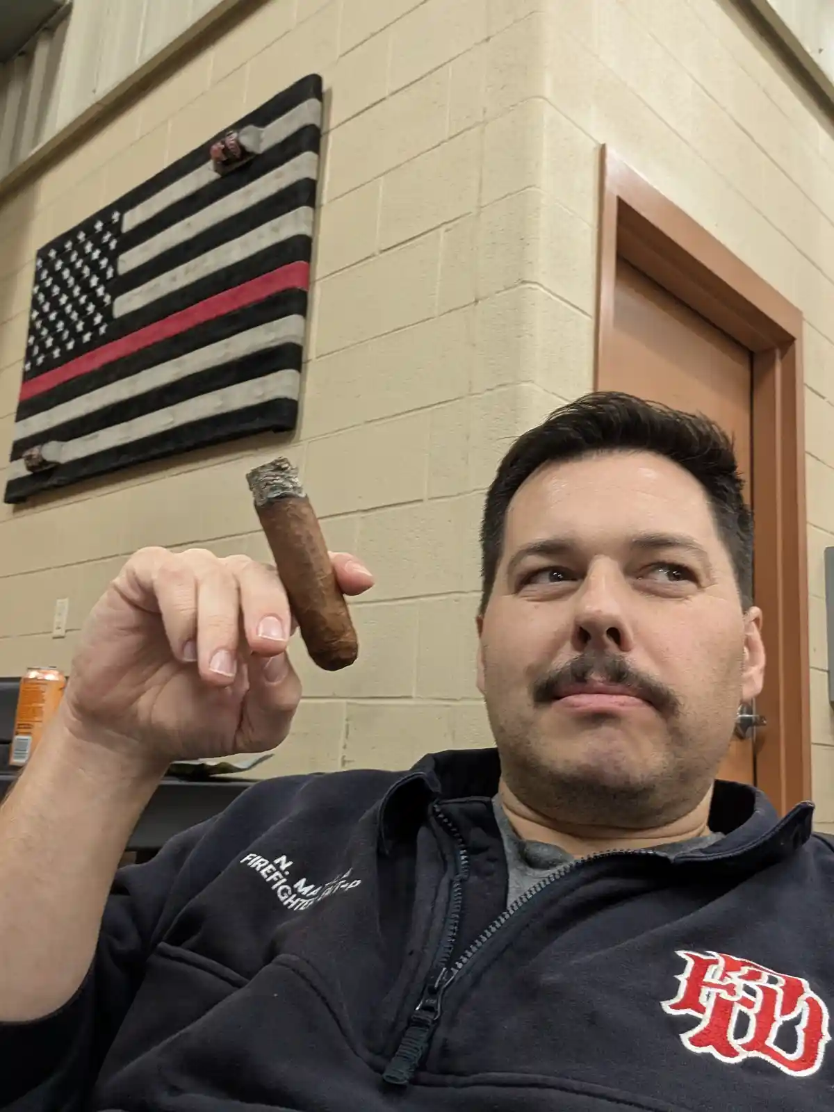 A man in a dark zip-up jacket with 'N. MAINE FIREFIGHTER' and a red 'FD' logo holds a lit cigar while looking upward with a…