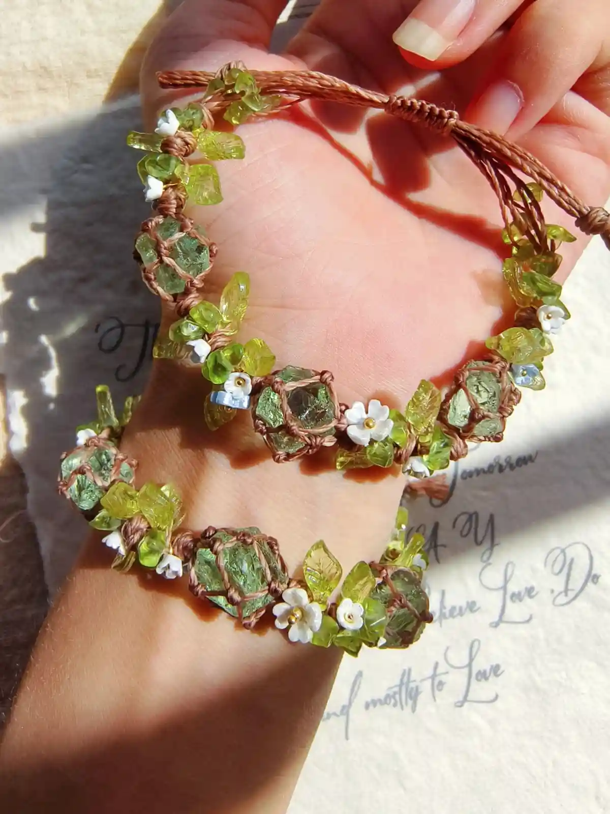A hand holds a handmade macrame bracelet with green gemstones and white flowers, set against a backdrop of a book with…