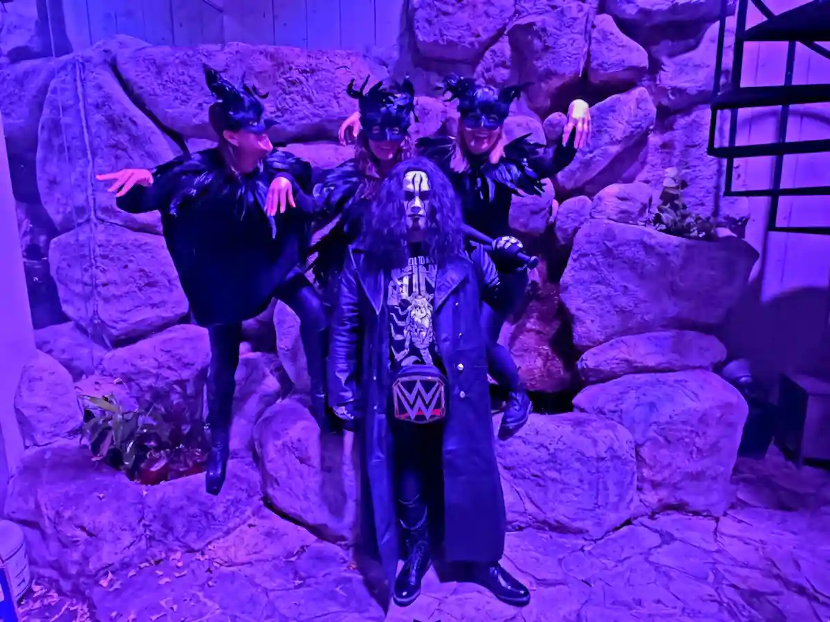 Four people in black, feathered, horned costumes with masks pose behind a central figure in a long coat and wrestling mask…