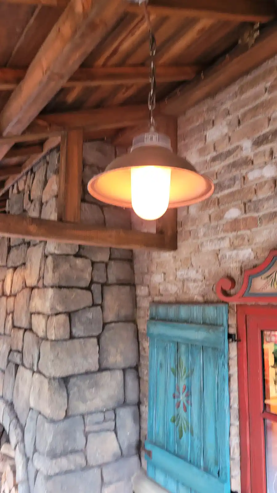 A warmly lit, vintage-style pendant lamp hangs from a wooden beam in a rustic setting with stone walls and a turquoise…