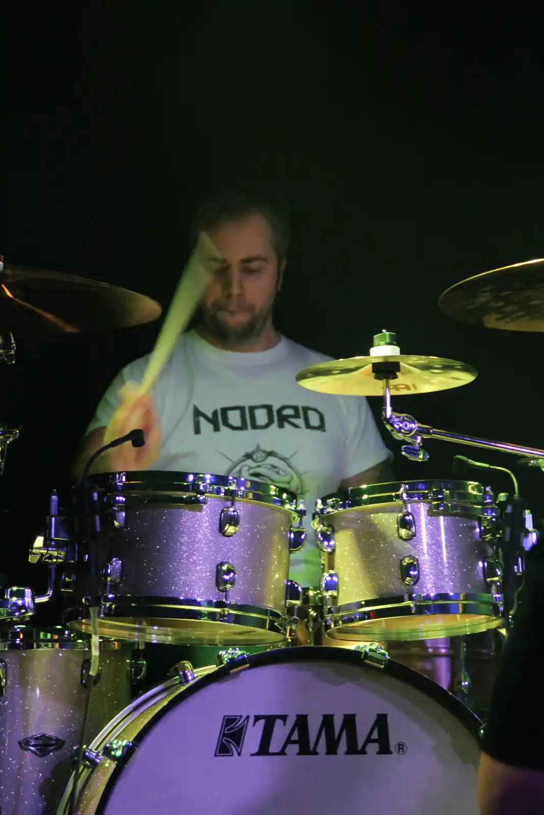A drummer in a white t-shirt with the word 'NODRD' printed on it is playing a glittery purple Tama drum kit under stage…