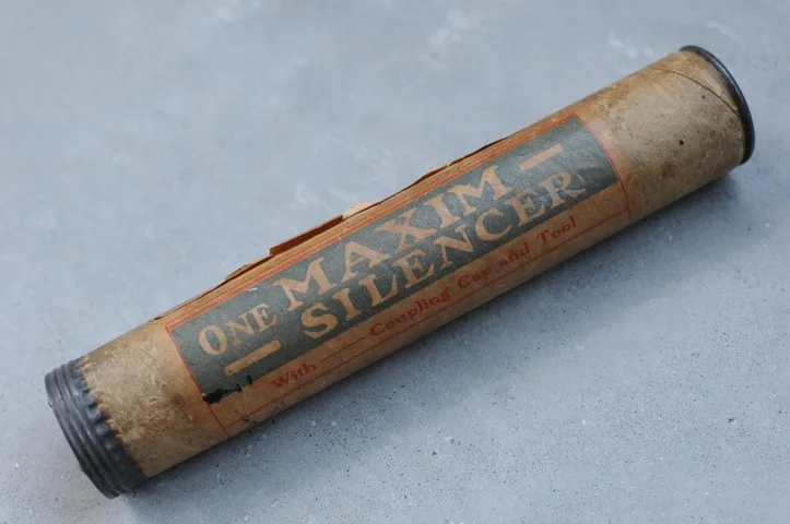A weathered, cylindrical cardboard package lies on a gray surface, labeled in faded red and black text: 'ONE MAXIM SILENCER…