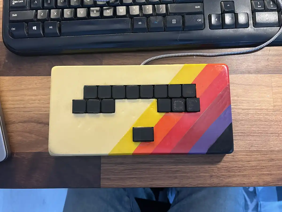 A small, custom-built keyboard with a rainbow-colored case and black keycaps sits on a wooden desk, positioned in front of a…