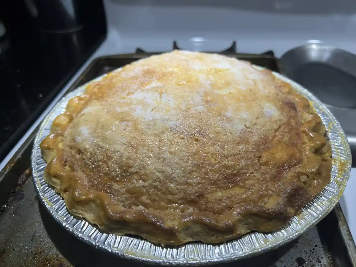 A freshly baked pie in a foil pan sits on a stovetop, its crust golden brown and slightly domed, with sugar crystals visible…