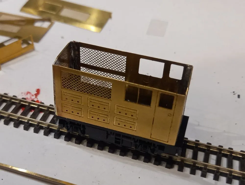 A brass model train car sits on a miniature track, its body showing detailed panel lines and mesh windows, with some parts…