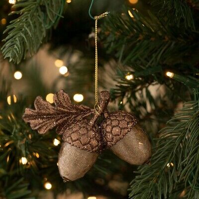 a glittery, brown decorative ornament shaped like two acorns with a leaf, hanging by a gold string from a green pine tree…