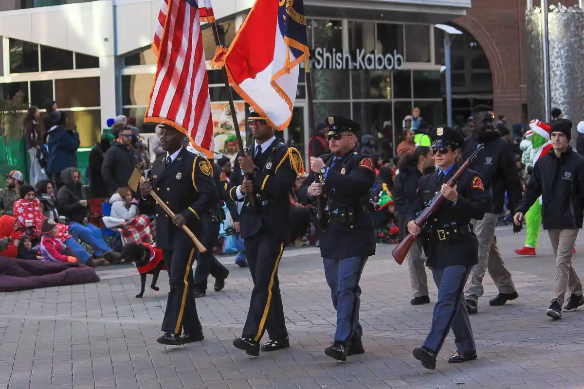A group of uniformed police officers in formal dress marching in a parade, carrying flags and a rifle. One officer holds an…