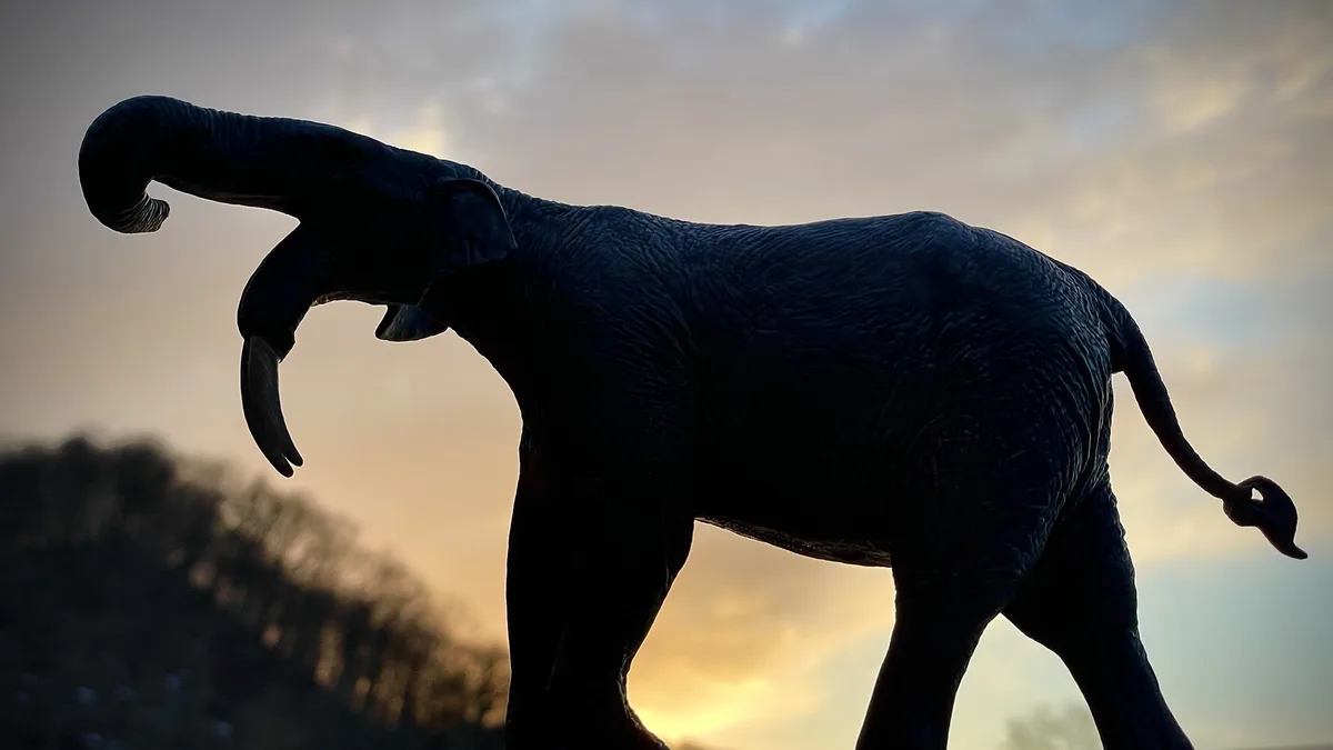 A silhouette of an elephant statue against a sunset sky, visibly featuring two trunks—one curled upward and one hanging…