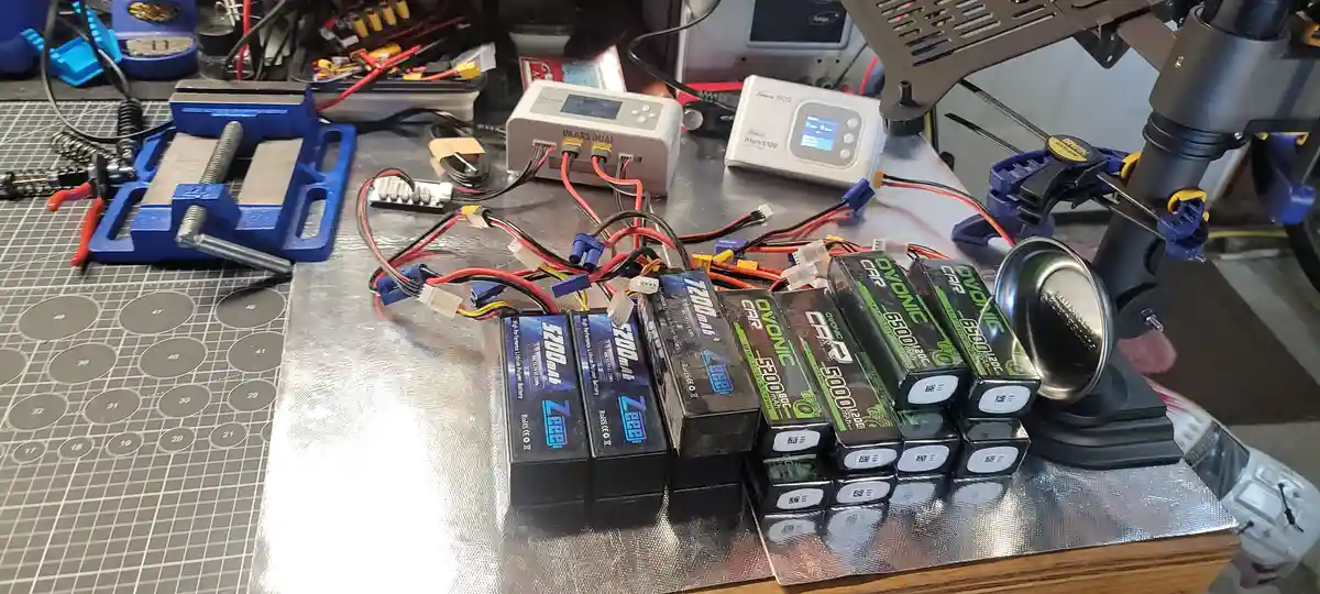 A cluttered workbench is filled with multiple lithium polymer batteries of various sizes and brands, tangled red and black…