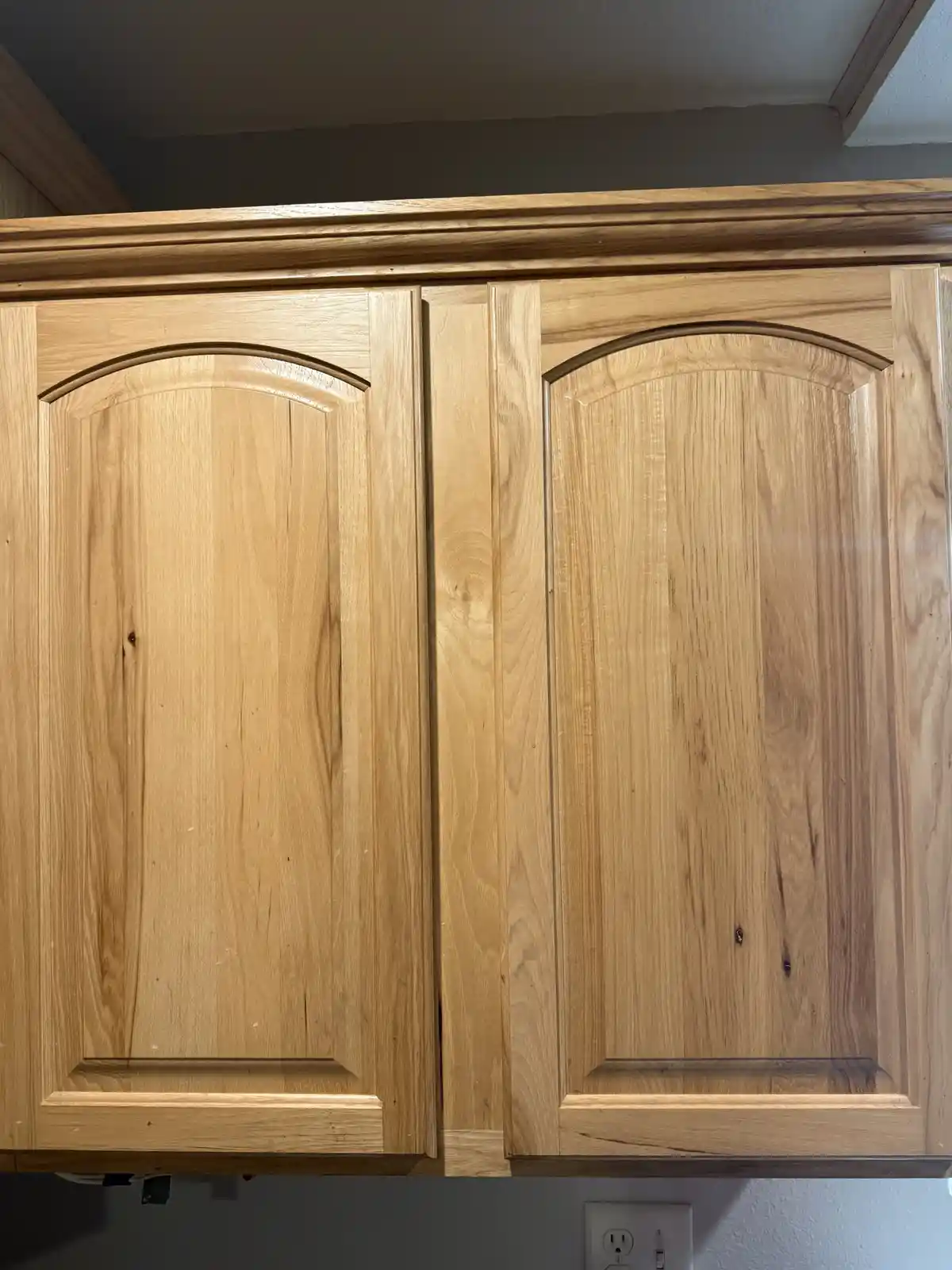 A close-up photo shows two light-colored wooden kitchen cabinets with arched tops and visible wood grain. The cabinets are…