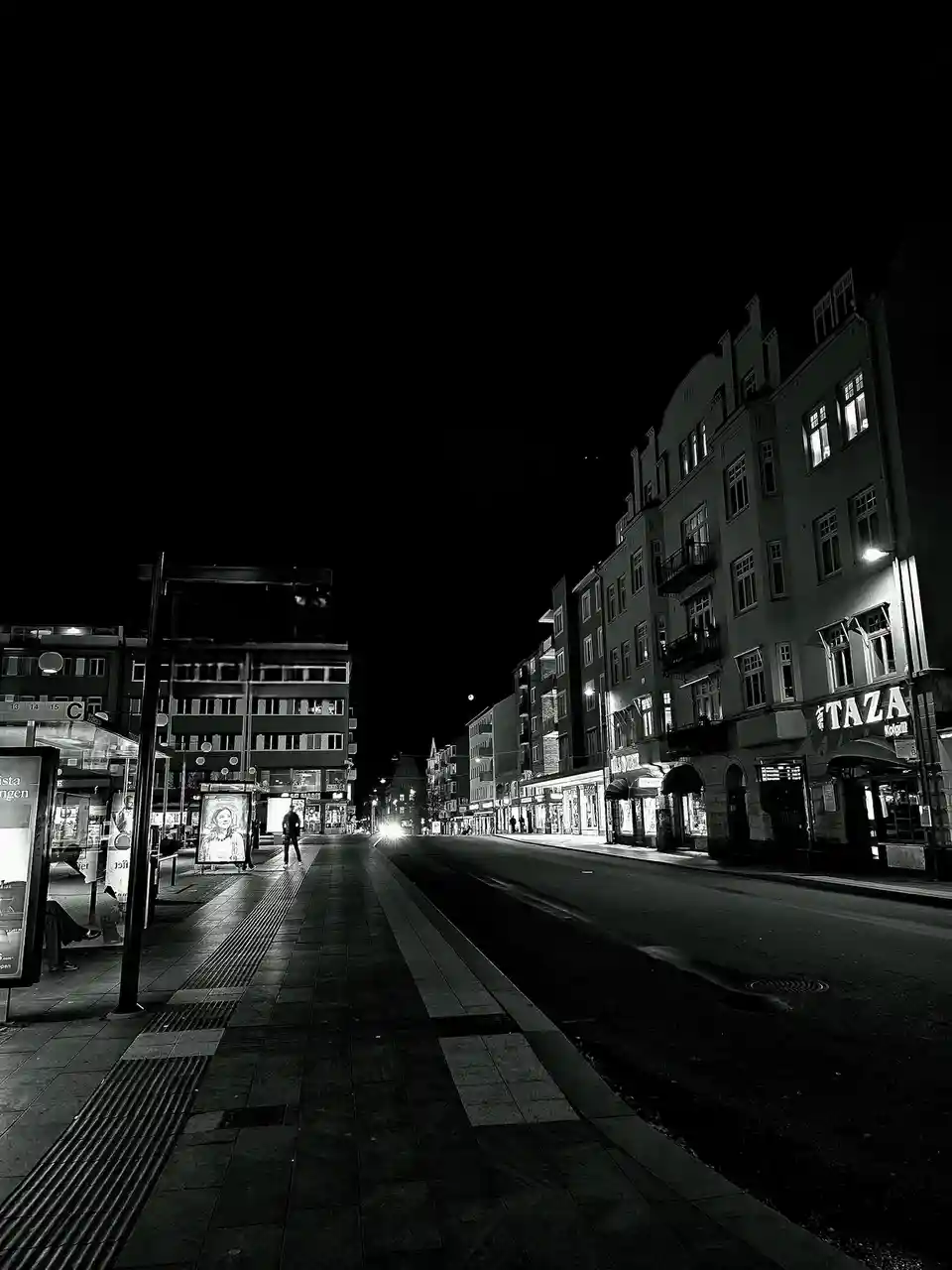 A black and white nighttime street scene with a lone person standing on a sidewalk near a bus stop, illuminated by…