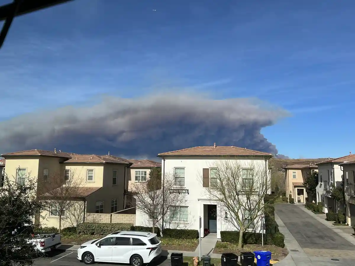 A suburban neighborhood with houses and parked cars under a clear blue sky, but a massive dark smoke plume from a wildfire…