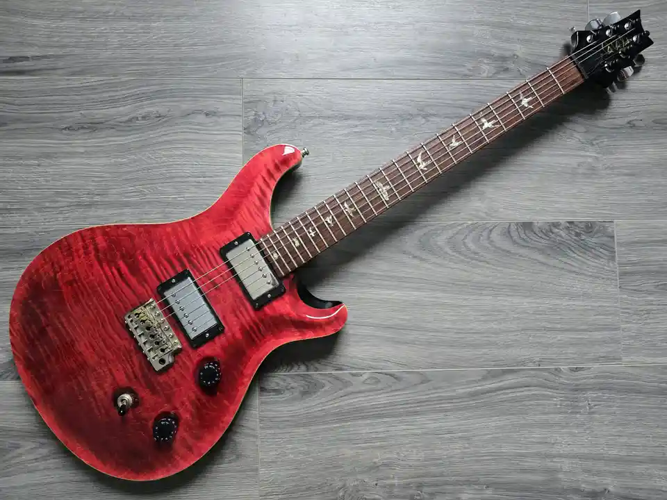 A red electric guitar with bird-shaped inlays on the fretboard lies diagonally on a gray wood-patterned floor. The overlaid…
