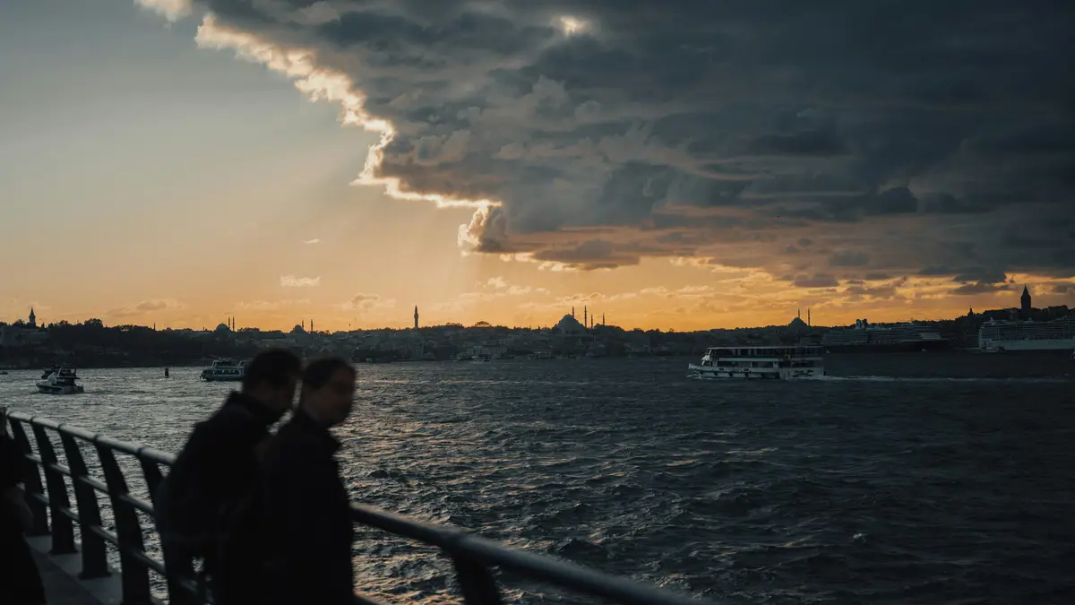 Vibrant hues of orange and pink illuminate the Bosphorus, casting a warm glow on silhouetted figures and boats, as dramatic…