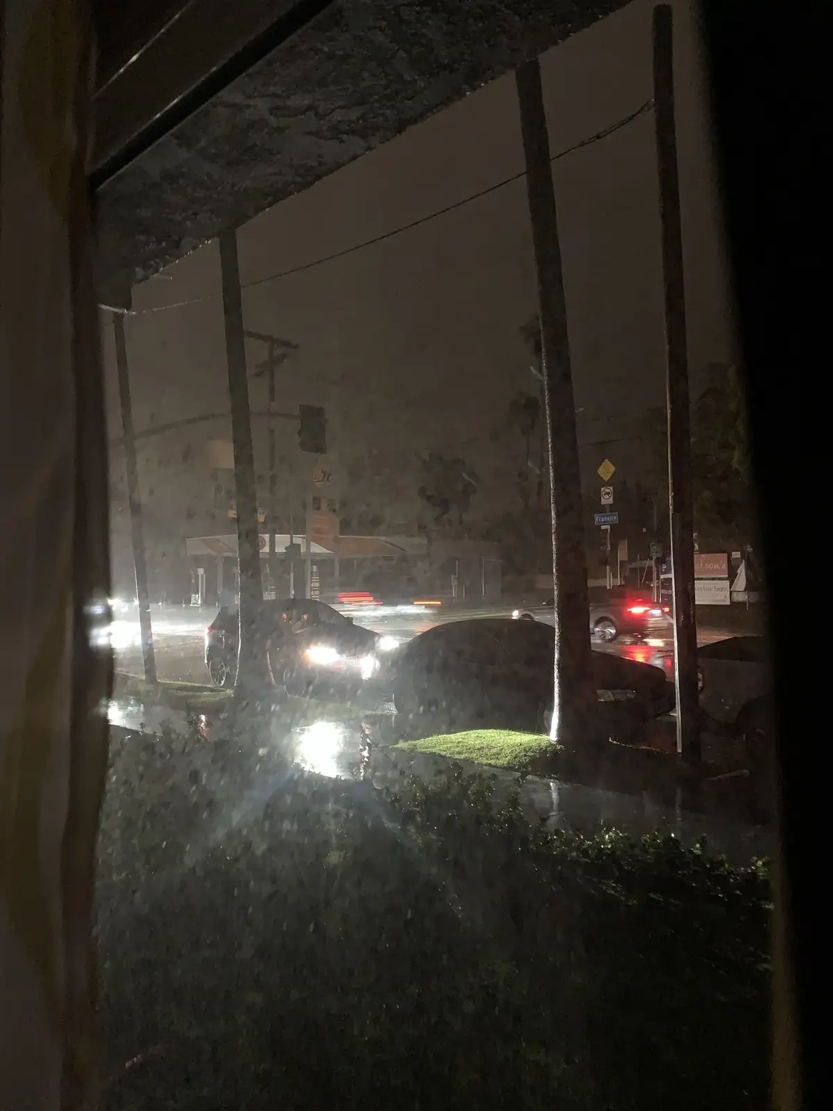 A nighttime view from inside a building looking out through a rain-splattered window at a street with cars and streetlights…