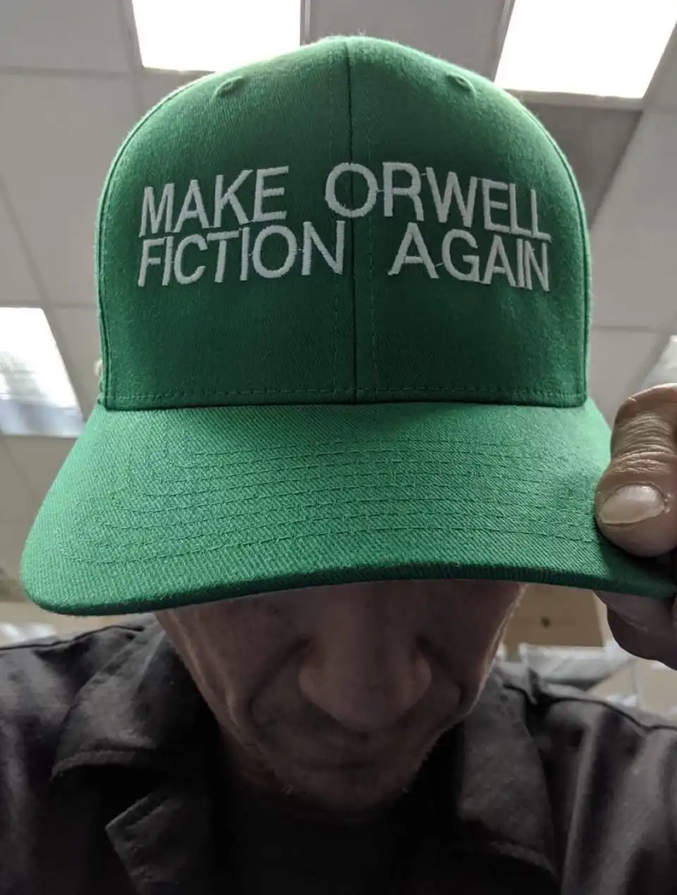 A close-up photo shows a person holding a green baseball cap with white embroidered text that reads 'MAKE ORWELL FICTION…