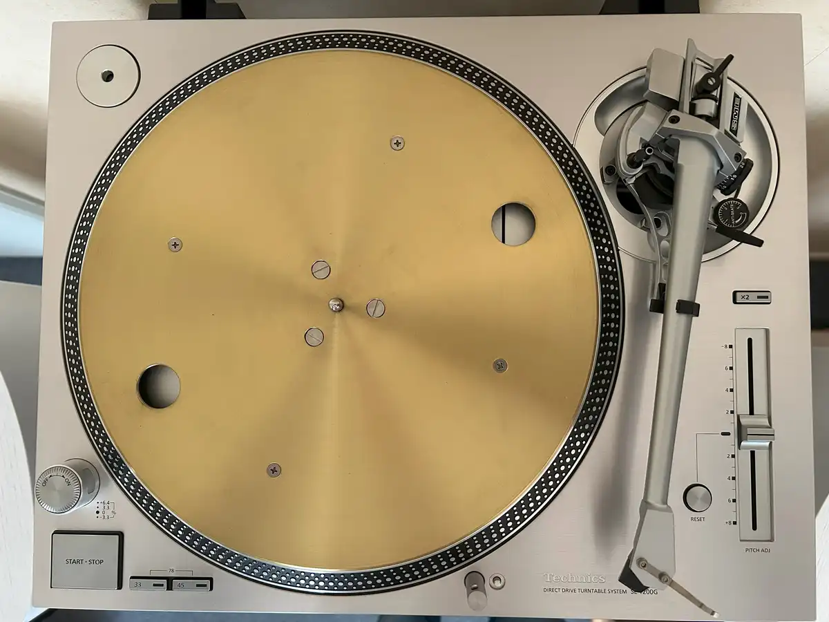 A top-down photo of a silver Technics SL-1200G direct drive turntable with a gold-colored platter, no record on it, and the…
